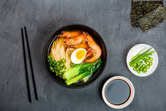 亚洲面汤、虾仁拉面、蔬菜和鸡蛋在灰色混凝土背景的黑色碗中。 平躺在地上,俯瞰,模拟,头顶. 健康食品概念