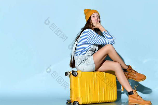 穿牛仔裤、全裙、条纹汗衫、<strong>靴子</strong>和帽子的少女。 她坐在黄色的手提箱上，蓝色的背景。 全长