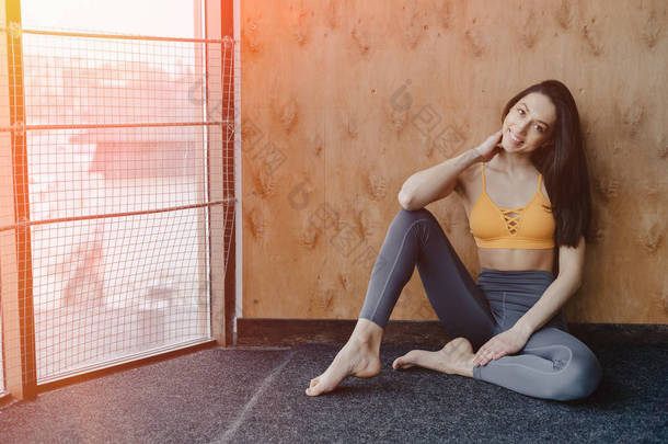 年轻迷人的健身女孩坐在地板上靠近窗户的木墙背景下, 在瑜伽课上休息