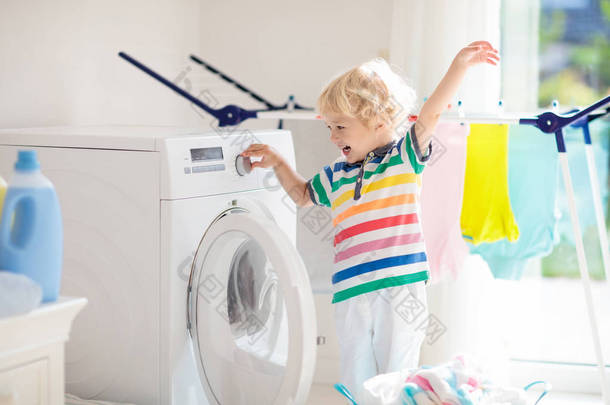 儿童在洗衣房内设有洗衣机或滚筒烘干机。<strong>帮助</strong>家庭琐事的孩子。现代家庭设备和洗涤洗涤剂在白色阳光的家。烘干机架清洗洗衣服. 