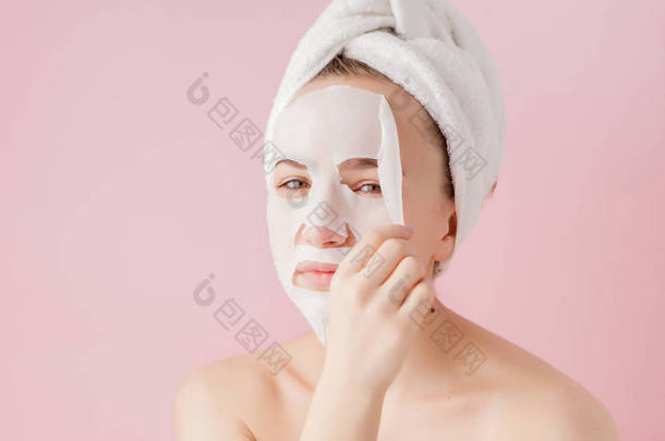 美丽的年轻女子正在涂上一个化妆品组织面膜的脸上的粉红色的背景。医疗和美容护理及技术理念