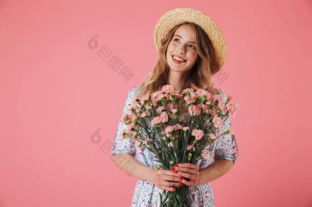 一个欢快的年轻女子在夏季礼服和草帽捧着康乃馨花束和看着远离粉红色背景的照片