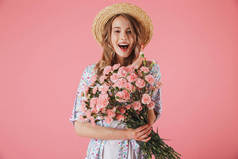 一个兴奋的年轻女子在夏季礼服和草帽持有康乃馨花束隔离粉红色背景的肖像