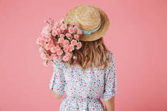 后视图的年轻女子在夏季礼服和稻草帽持有康乃馨花束隔离粉红色背景的肖像