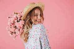 关闭一个可爱的年轻女子在夏季礼服和草帽捧着康乃馨花束, 看着她的肩膀被孤立的粉红色背景的肖像