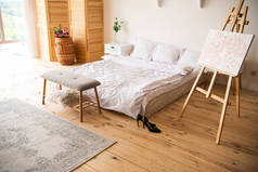 卧室配有白色毯子和枕头、画架、床头柜、地毯、房间铺垫和木地板上的黑色高跟鞋