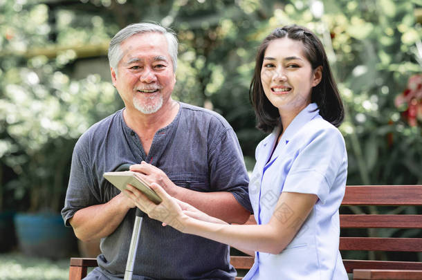 护士与病人坐在板凳上一起看片剂。亚裔老人和年轻女子坐在一起说话。放松, 快乐的微笑心情.