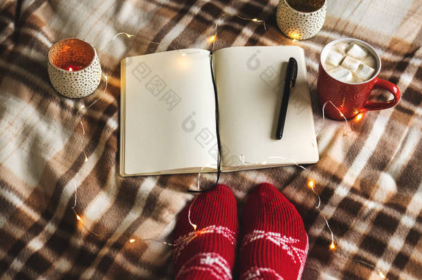 秋季和冬季背景与笔记本, 钢笔, 花环, 蜡烛, 女人的脚在针织红袜, 一杯热巧克力与棉花糖的格子。工作理念, 在温馨的家中制定计划.
