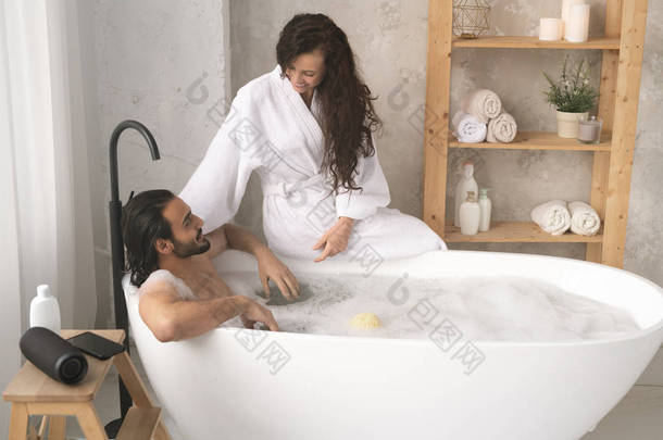 穿着白色浴衣的快乐的年轻女人坐在浴缸上，和丈夫聊天，享受着泡沫浴