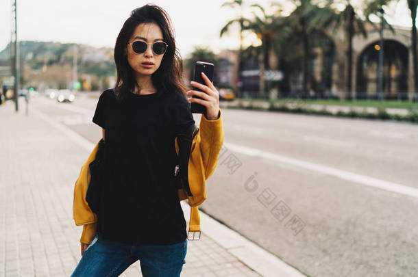 时尚的亚洲妇女与长的深色头发在太阳镜穿着黑色 t恤衫和黄色皮夹克在一个模糊的城市背景下采取自拍的手机.
