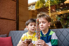两个弟弟男孩在户外用巧克力和五颜六色的糖洒着松饼，吃着香甜的水果。 不健康美味甜食概念