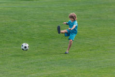 穿着运动服的卷发男孩在绿草地上踢足球 