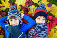 两个小男孩穿着五颜六色的休闲装躺在秋天的树叶里。在温暖的日子里，快乐的兄弟姐妹们在秋天的公园里玩得开心。有枫叶的健康儿童围巾和帽子