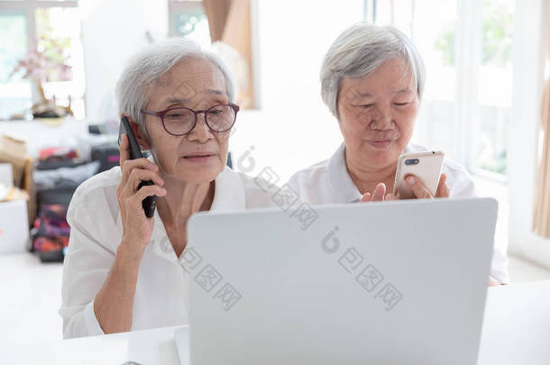亚洲老妇人和朋友带着<strong>笔记本电脑</strong>，快乐微笑的老人一边看一些有趣的事情，一边拿着电话，谈论家里的金融业务，科技概念 