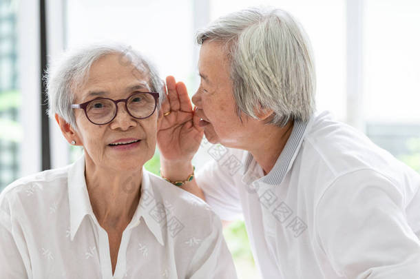 亚洲老妇人手牵着嘴说搞笑，耳边八卦的朋友，说话在老年妇女的耳朵和近脸，有听力<strong>困难</strong>，听力障碍，听力受损，听力受损的老人