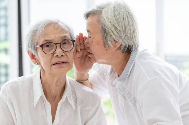 亚洲老妇人手牵着嘴说搞笑，耳边八卦的朋友，说话在老年妇女的耳朵和近脸，有听力困难，听力障碍，听力受损，听力受损的老人