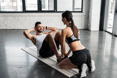 运动的女孩帮助快乐的年轻人做腹肌运动在瑜伽垫在健身房