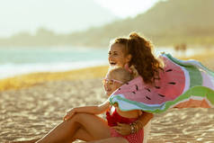 快乐的现代母亲和孩子穿着海滩, 拿着西瓜毛巾坐在海边, 晚上玩得很开心。最小到没有人群和平。戴着太阳镜的金发孩子.