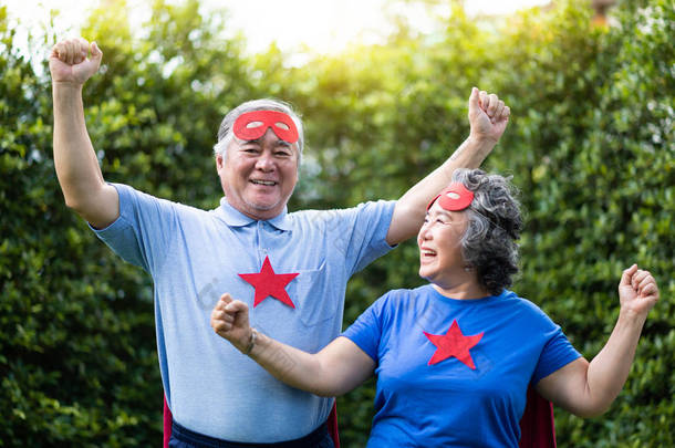亚洲资深夫妇在超级英雄服装放松和<strong>庆祝</strong>与<strong>胜利</strong>。老年人戴着红色的面具和蓝色的衬衫, 明星们玩得很开心, 在公园的户外一起微笑.