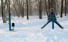 女人在蓝色羽绒服与胡德是做腿演习在模拟器在冬季公园前视图