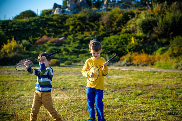 两个活跃的学龄前男孩夏天在室外场地打羽毛球。孩子们打网球。儿童学校体育活动。儿童运动员的网球拍和羽毛球运动。朋友们在一块温暖的田野里快乐.