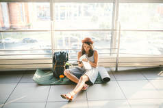 等候, 延迟运输在机场或火车站的终端。穿着和帽子的年轻白种女人坐在旅游毯上, 背包靠近窗户到房间, 看着旅游地图。..