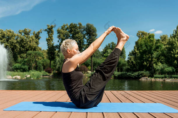 在公园附近的河边瑜伽垫上练习瑜伽和伸展腿的妇女的侧面视图