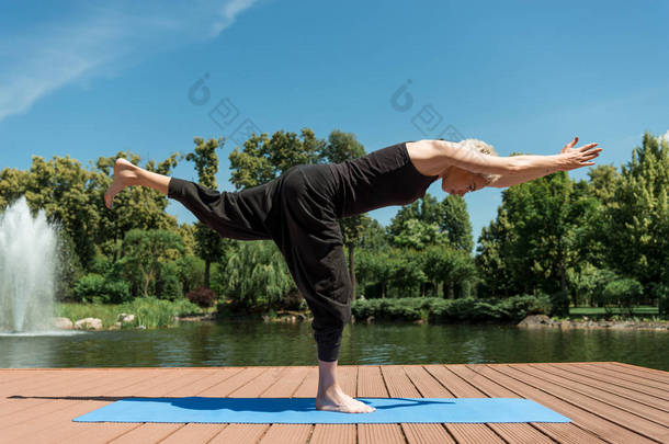 女子练习瑜伽的侧面视图 iii (Virabhadrasana iii) 在公园河边的瑜伽垫上摆姿势