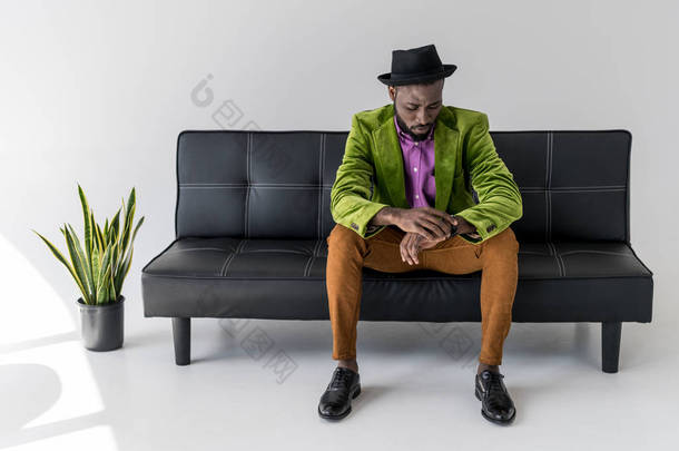 非洲裔美国人时尚男子在帽子检查时间, 而坐在黑色沙发上