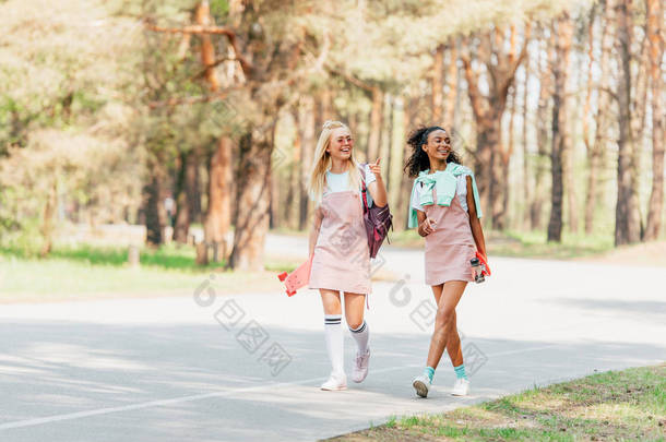 两个微笑的多元文化朋友拿着便士板，走在路上的全长视图