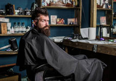 戴着黑斗篷的大胡子坐在镜前的美发椅上。男子与胡子客户的时髦理发店。理发店概念。时髦与胡子等待理发和理发