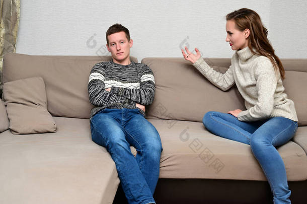 家庭冲突的概念。 一<strong>个</strong>不快乐的年轻家庭的画像：愤怒的黑发女孩和不高兴的家伙穿着毛衣躺在房间里的沙发上。 不满意的妻子责备丈夫.