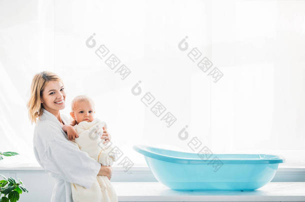 快乐的母亲在浴袍上的侧面视图在塑料婴儿浴缸附近携带可爱的孩子裹着毛巾