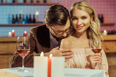 微笑的女孩用酒杯看着相机, 而她的男朋友亲吻她的肩膀在餐桌上的餐厅 