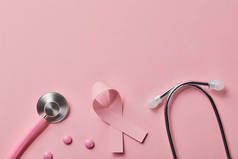 粉红金属听诊器、乳腺癌丝带和浅粉色背景上的三片药片的顶视图