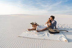 一对年轻夫妇抱在怀里，坐在毛毯上，沙滩上挂着一筐水果和声吉他