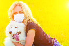美丽的卷曲金发碧眼的快乐的年轻女子穿着牛仔短裤，戴着面具，在夏日的夕阳西下的田野里，拥抱着一只毛茸茸的可爱的萨莫伊犬。宠物和女主人