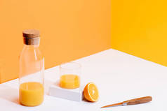 新的橙汁在玻璃杯中，瓶子在成熟的橙子附近，刀片在橙色背景的白色表面