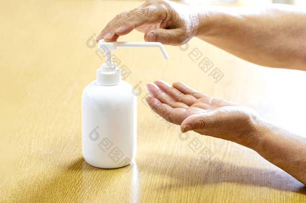 一位老年人用塑料喷雾瓶中的防腐剂喷洒双手。手与瓶子在木制背景的特写。流行病期间的卫生治疗.