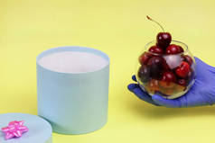 成熟的甜樱桃放在玻璃碗里，手戴蓝色手套，背景是黄色的。脱毛，圆形，礼品盒。复制空间.新鲜水果、假日、安全、检疫、珊瑚的概念