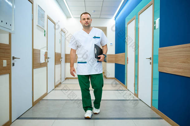 穿着制服的友善的男医生在诊所走廊里拿着笔记本。医院专业医疗专家、喉科或耳鼻喉科专家、妇科医生或乳腺专家、外科医生