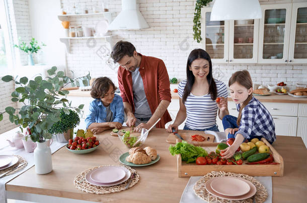准备沙拉。年轻漂亮的一家人在家里的现代化厨房里一起做饭.爸爸妈妈教两个小孩如何切新鲜蔬菜