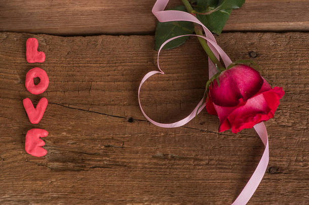 红<strong>玫瑰</strong>一种红<strong>玫瑰</strong>花，有粉红色的缎带，<strong>心形</strong>环绕着，爱这个词从塑料线中伸出，放在老式的木制表面，顶部的景色上。带有空间的浪漫明信片。浪漫的爱情告白