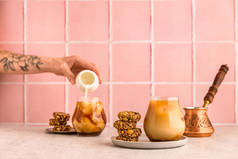 一杯冰镇咖啡，配上燕麦饼干。一个cezve 。一个女人的手，手上拿着花纹身，从一个白色的罐子里倒入牛奶。温暖的光线，明亮的图像，粉红的瓷砖背景