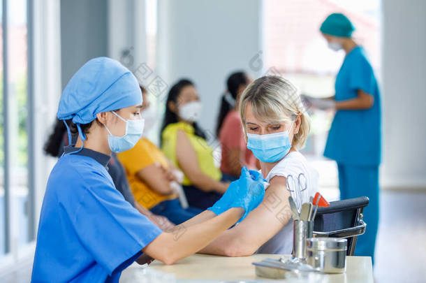 亚洲女医生头戴面罩，身穿蓝色医院制服，为白种人老年妇女注射疫苗，并配备设备，而其他病人则在模糊的背景下<strong>排队</strong>等候.