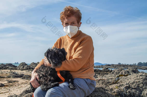 老女人带着她的狗在海滩上戴着医疗面具，这是新常态中的休闲观念