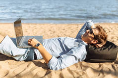 戴眼镜的年轻人, 躺在沙滩上, 在沙滩上的笔记本电脑上工作, 在海边工作, 度假,