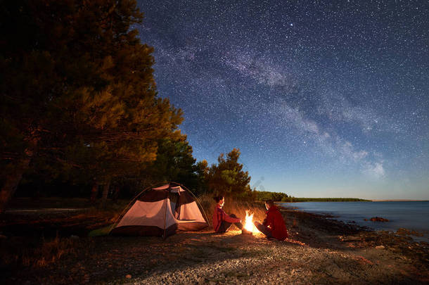 晚上在岸上露营。男子和女子徒步旅行者在帐篷前休息, 在黄昏的天空中充满了星星和银河的蓝色水和森林背景下的篝火。户外生活方式概念