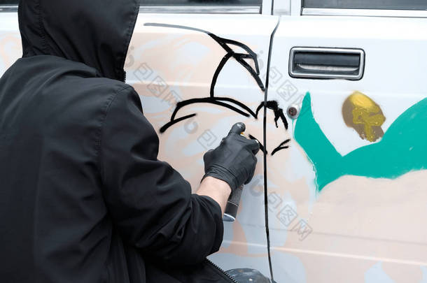 青年亚文化涂鸦。一个穿着黑色衣服和手套的家伙在一辆白色汽车上<strong>画</strong>了一个抽象的<strong>形象</strong>。破坏或艺术.