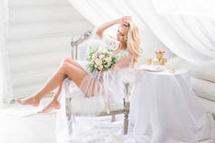 长腿金发的新娘带着一束温柔的花朵, 羞怯地微笑着, 她正坐在一张舒适的白色椅子上, 美丽的内饰。新娘的早晨。美术婚礼